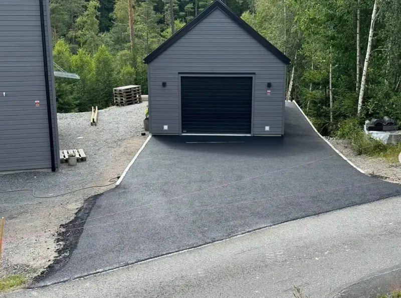 Nylagd asfaltsinfart som leder fram till ett mörkgrått garage, kantad mot grus och skog.