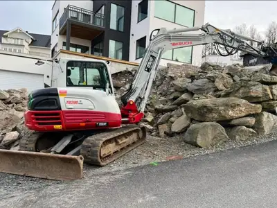 Grävmaskin märkt Takeuchi TB290 står vid uppschaktade stenmassor intill villabebyggelse.