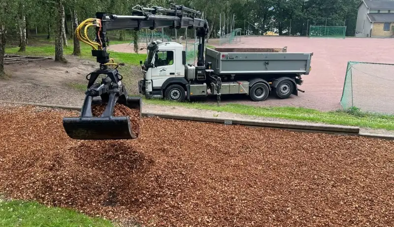 Kranbil med griptång lägger ut täckbark på en yta vid en idrottsplats.