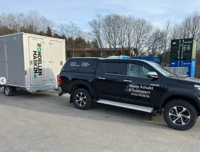 Svart pickup märkt Akam Schakt & Transport med kopplad släpvagn.
