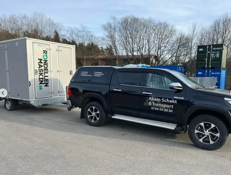 Svart pickup märkt Akam Schakt & Transport med kopplad släpvagn på en uppställningsplats.