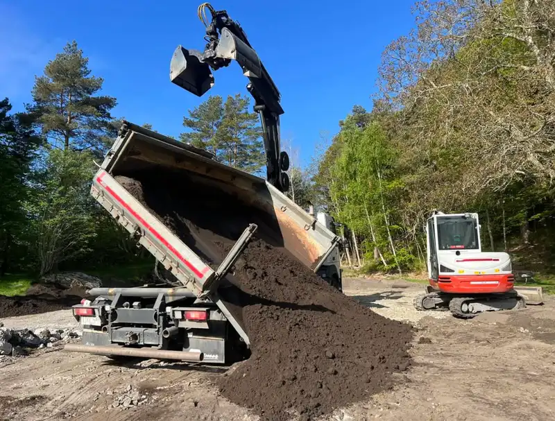 Lastbil med kranarm tippar av jordmassor, grävmaskin står bredvid på grusytan.